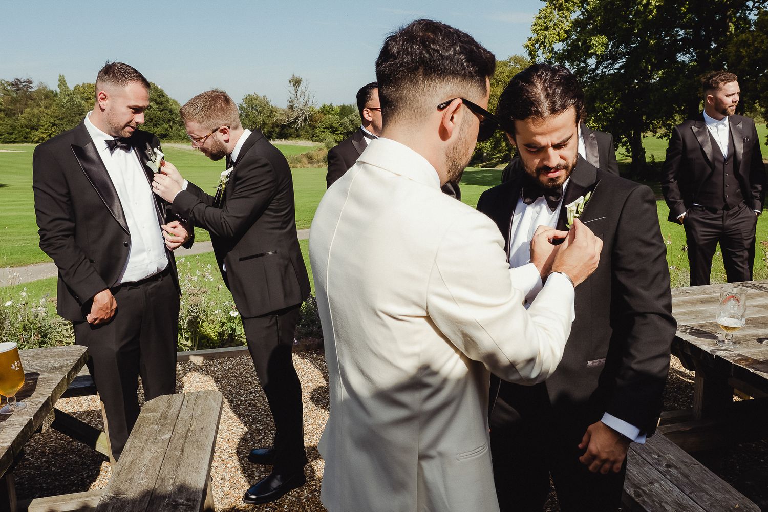Groomsmen in black suits gather outdoors on a sunny day for a candid wedding photo session.
