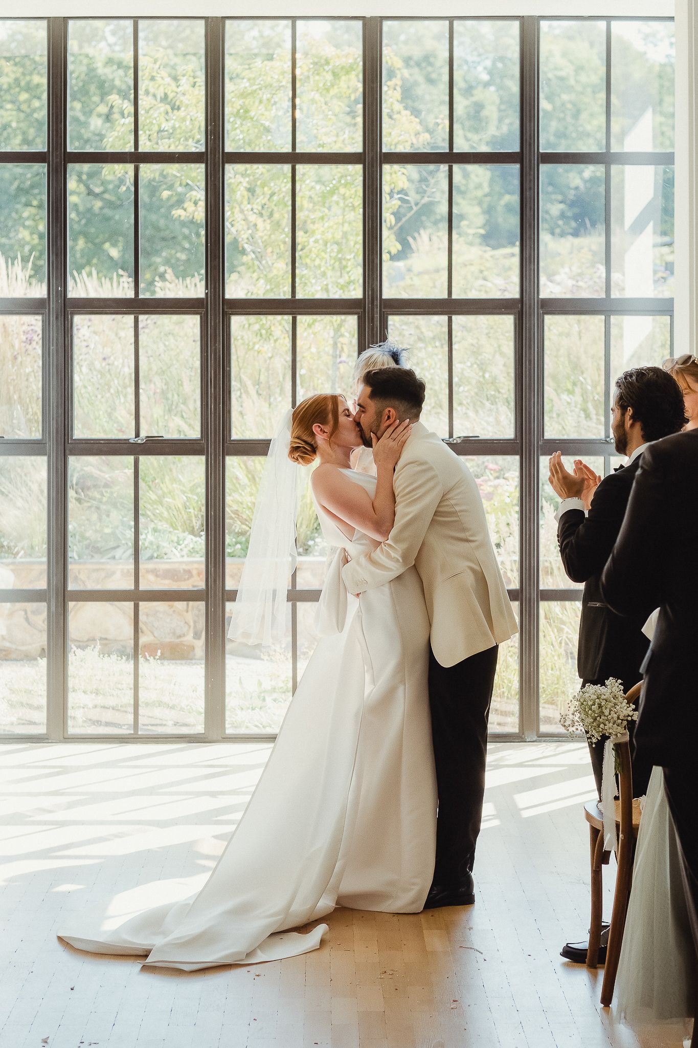 Elegant wedding couple shares their first kiss in a sun-filled room with floor-to-ceiling windows and natural light.