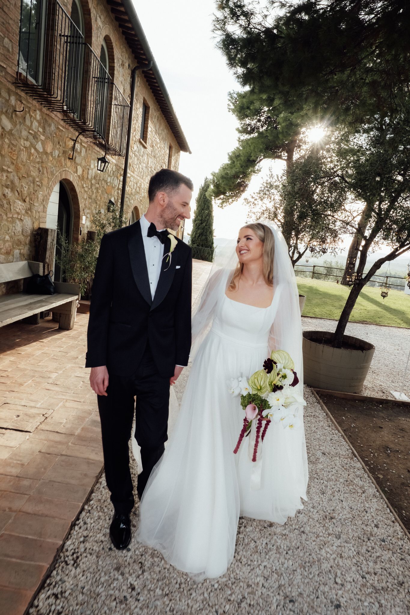 Smiling bride in a white gown and veil holding a bouquet walks beside a groom in a black tuxedo at a stone villa in Tuscany.