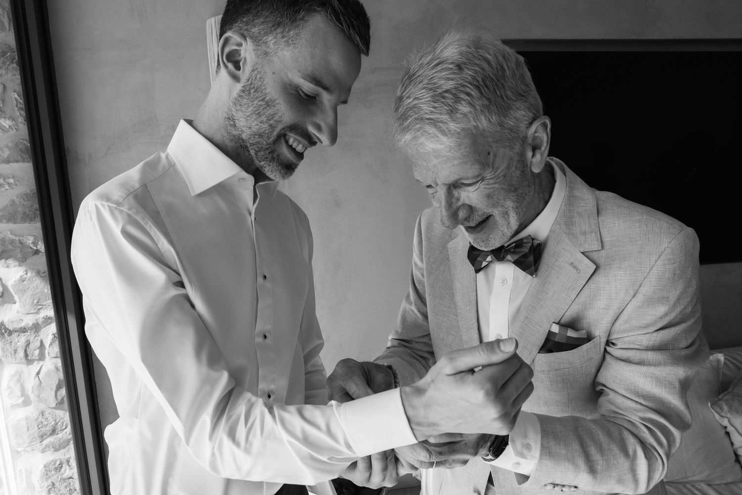Smiling groom in a white shirt helps an older man in a light suit and bow tie fasten his cufflink in a black and white wedding portrait.