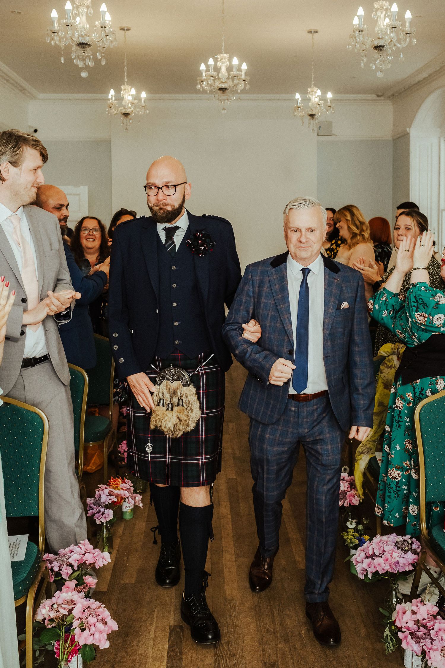 Two men in kilt and tartan suit walk arm in arm down the aisle between pink flower arrangements as wedding guests smile and applaud.