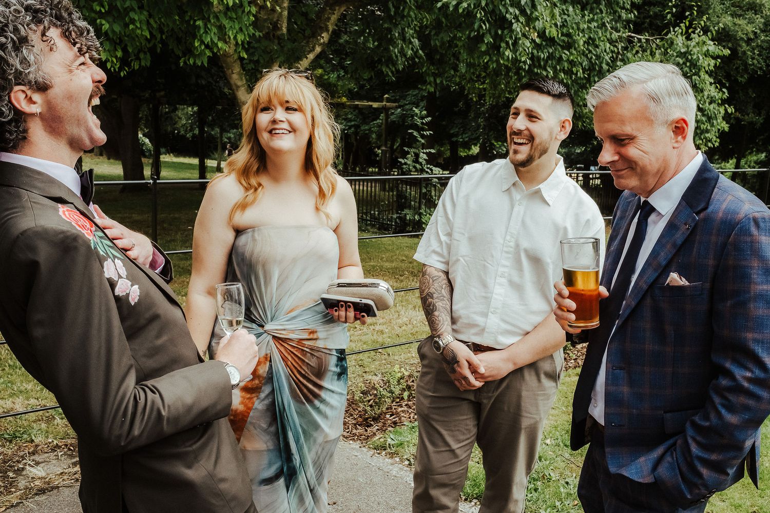 Four smiling guests chat and laugh outdoors at a wedding, holding drinks beneath green trees on a sunny day.