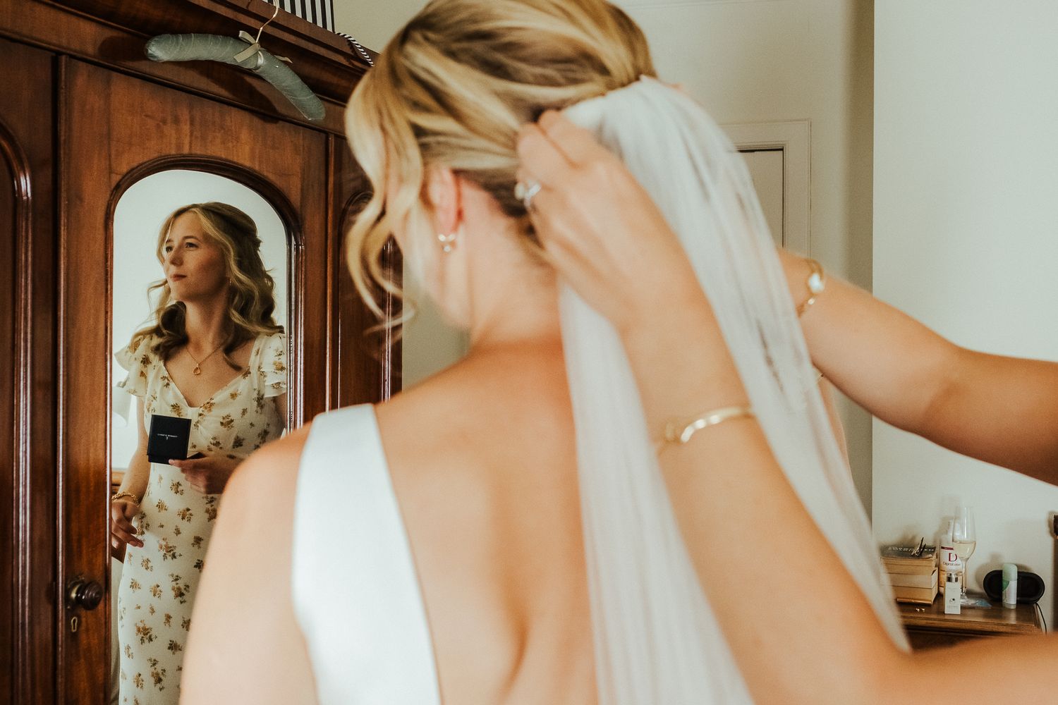 A bride adjusting her veil in front of an antique wooden mirror during wedding preparations.