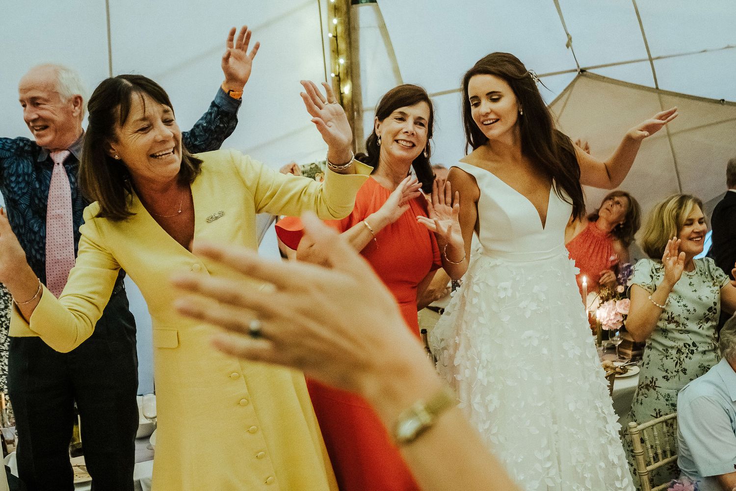 Energetic wedding party dances and celebrates under string lights at outdoor reception.