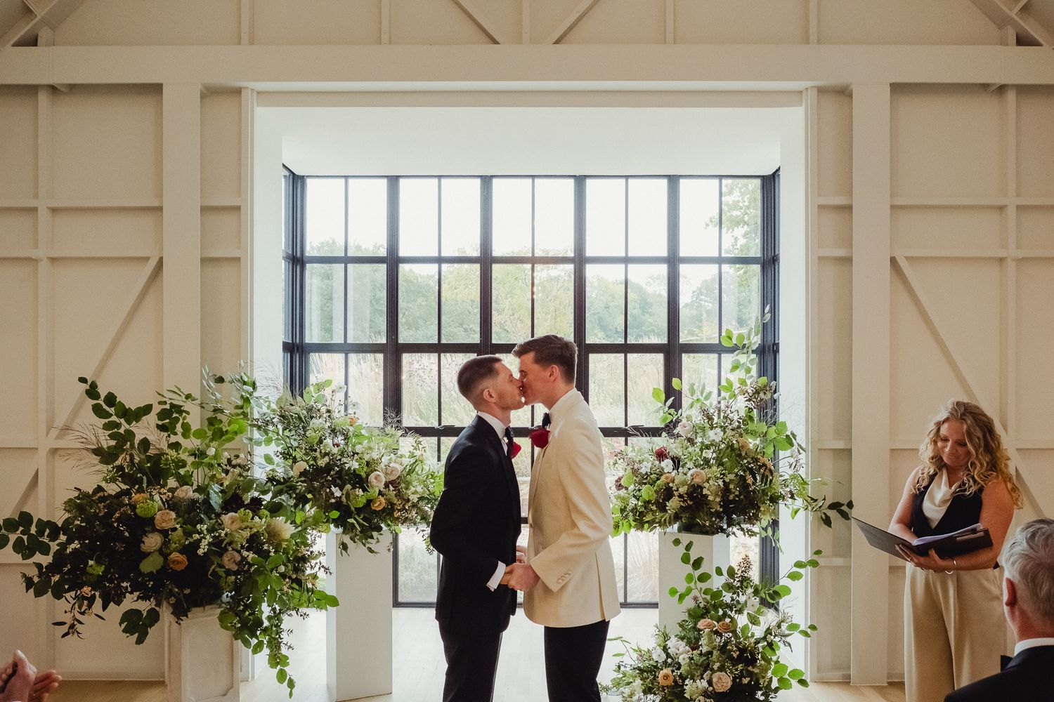 A couple shares a romantic moment at the altar during their wedding ceremony in a bright white barn venue with floral arrangements.