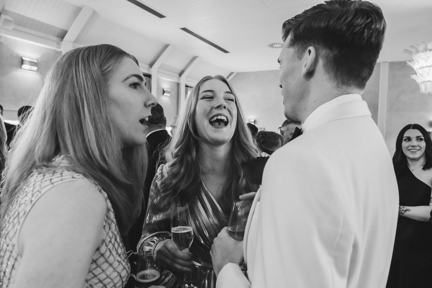 A black and white candid photo shows a group of people laughing and socializing at an indoor event.