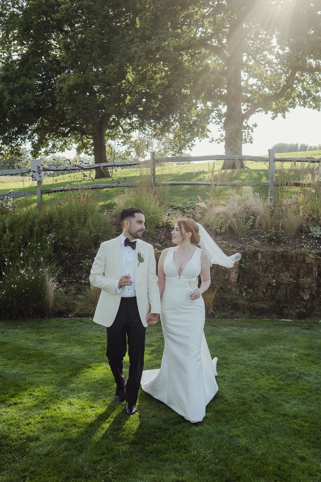 Newlyweds dance joyfully together on a sunlit lawn with trees and stone wall in the background during golden hour.
