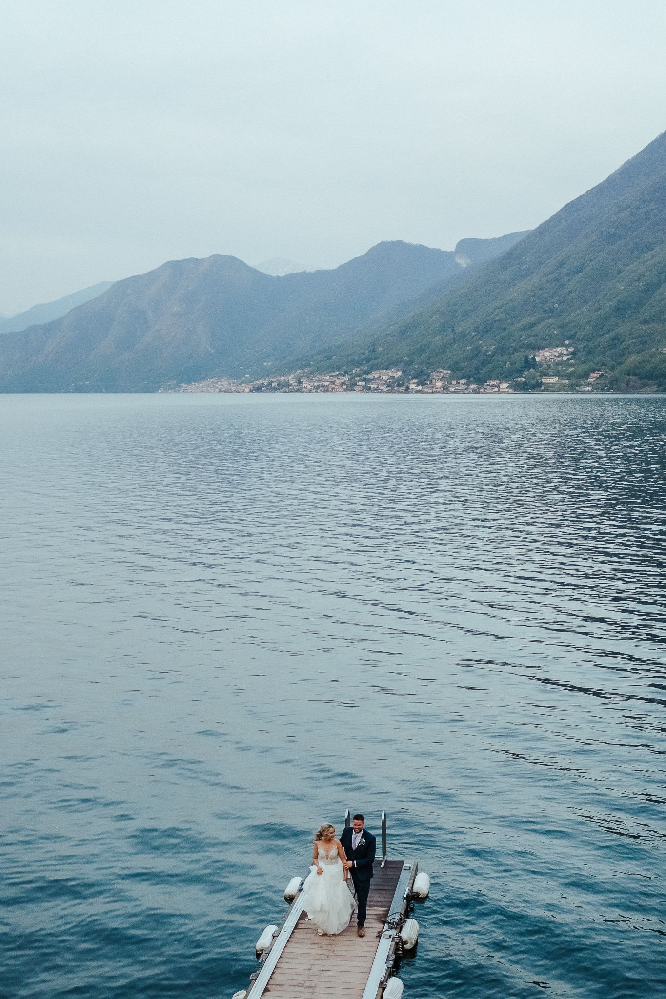 A couple sits at the end of a wooden pier overlooking a serene mountain lake surrounded by misty peaks.
