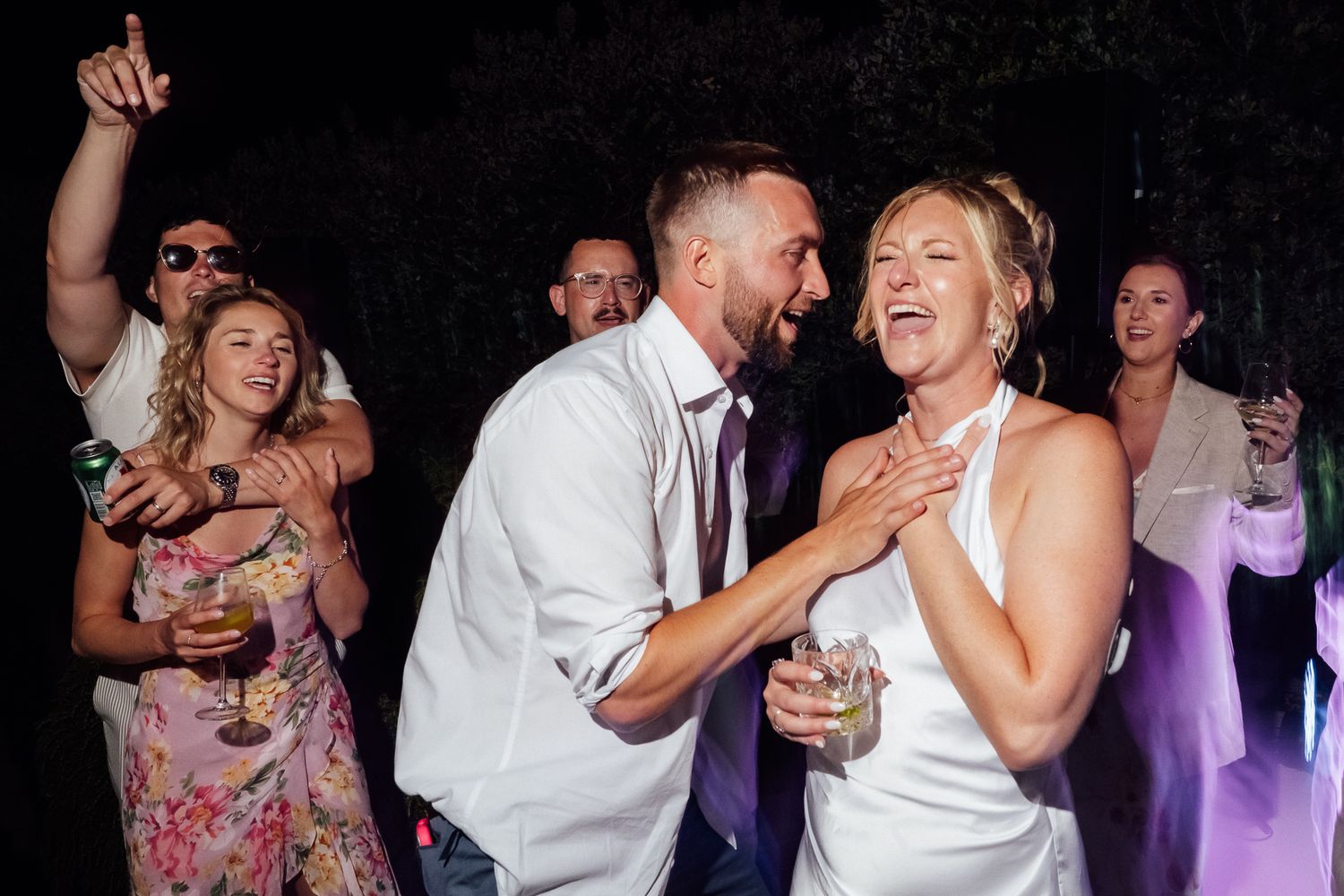 Joyful bride and groom dancing together at wedding reception with guests celebrating in background.