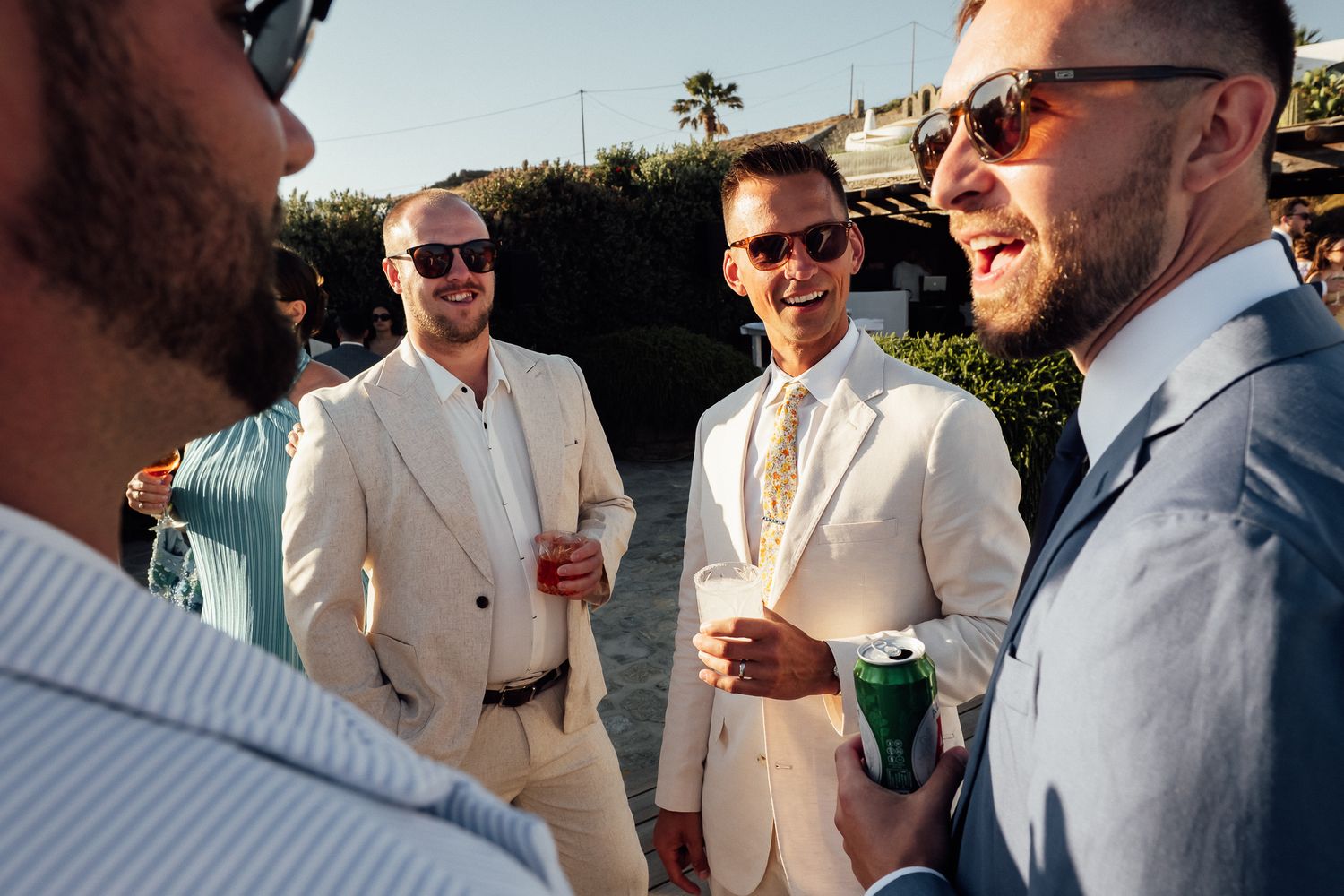 Groomsmen in beige suits and sunglasses laughing and drinking at an outdoor wedding reception.