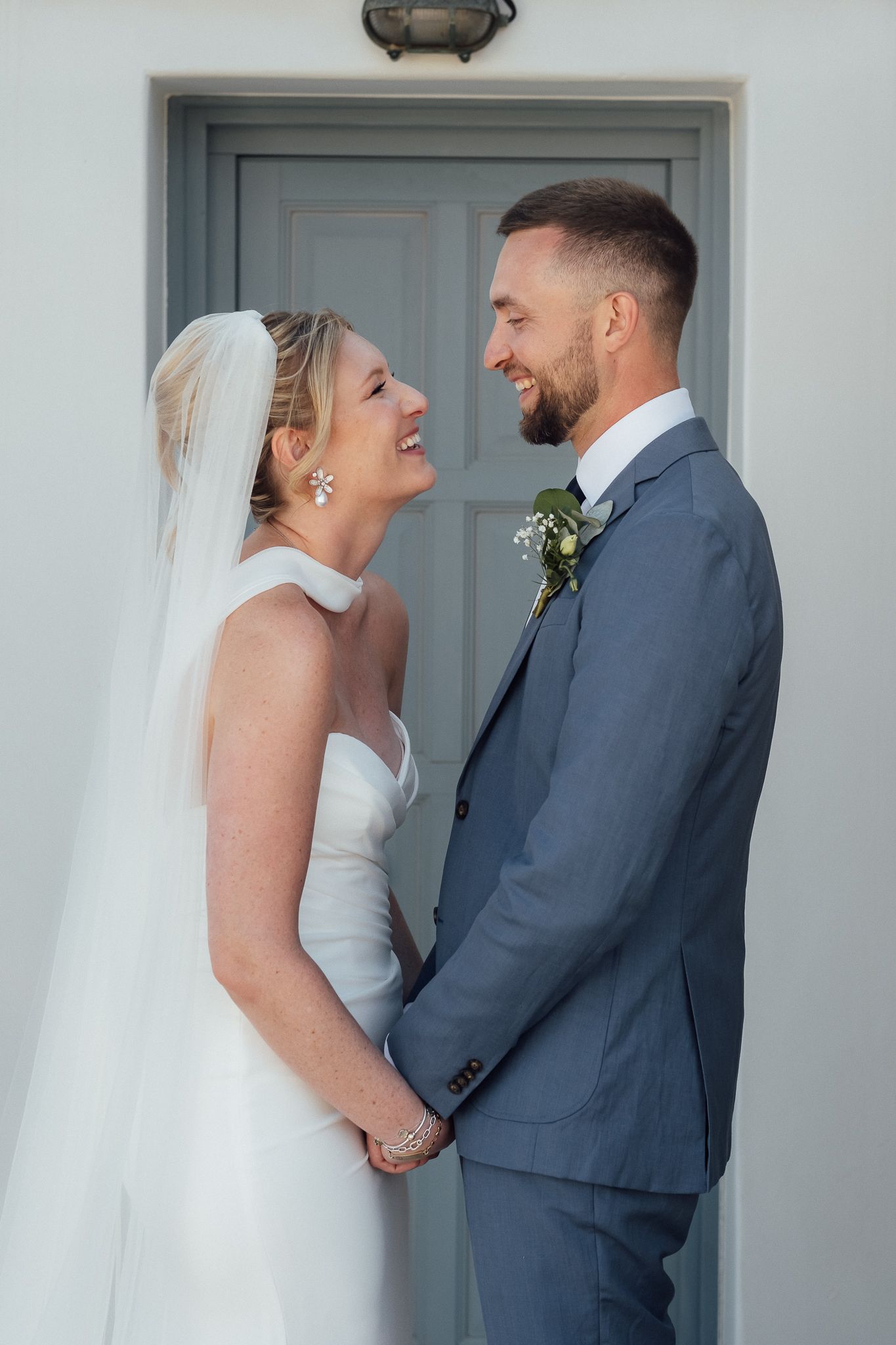 A smiling bride and groom hold hands in front of a gray door, dressed in a white gown and blue suit.