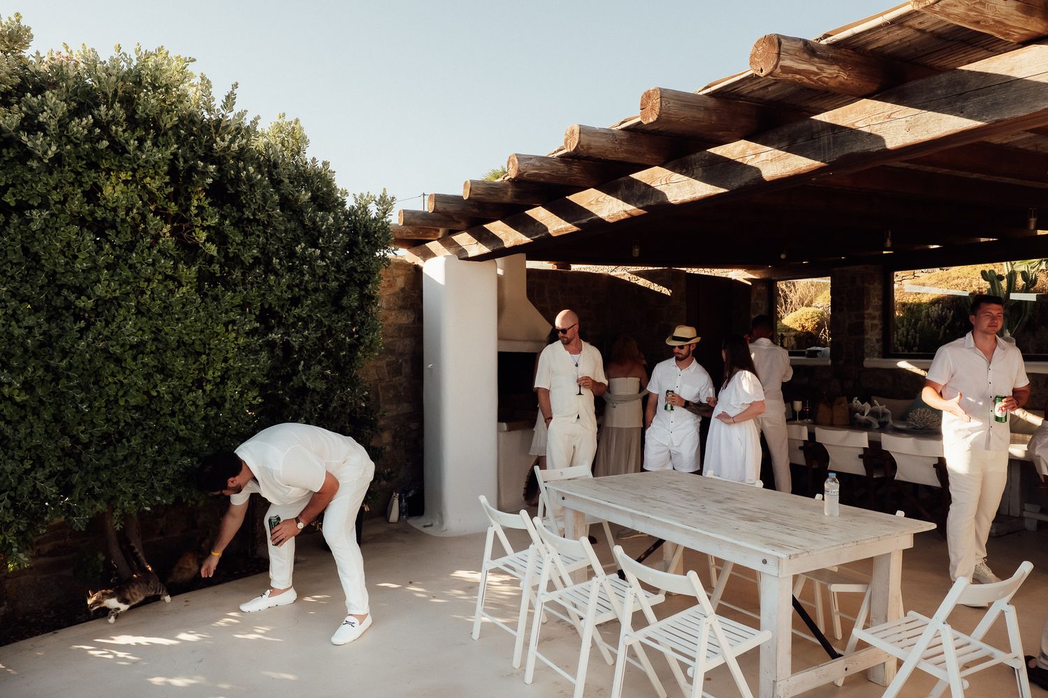 Staff in white uniforms prepare an outdoor dining area with white chairs and a wooden pergola in a sunny Mediterranean setting.