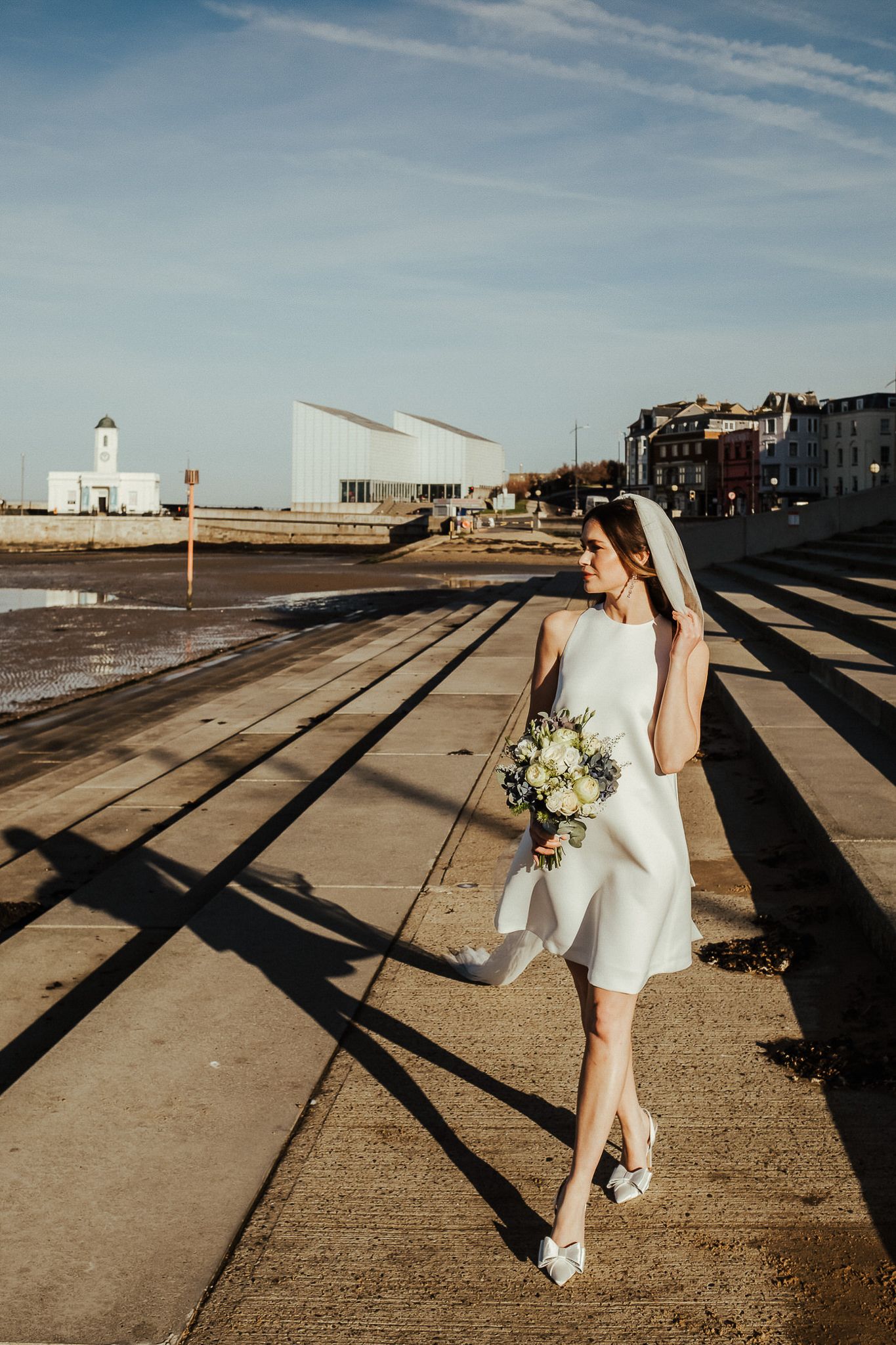 A romantic seaside photoshoot featuring a white dress and bouquet against a lighthouse and coastal backdrop.