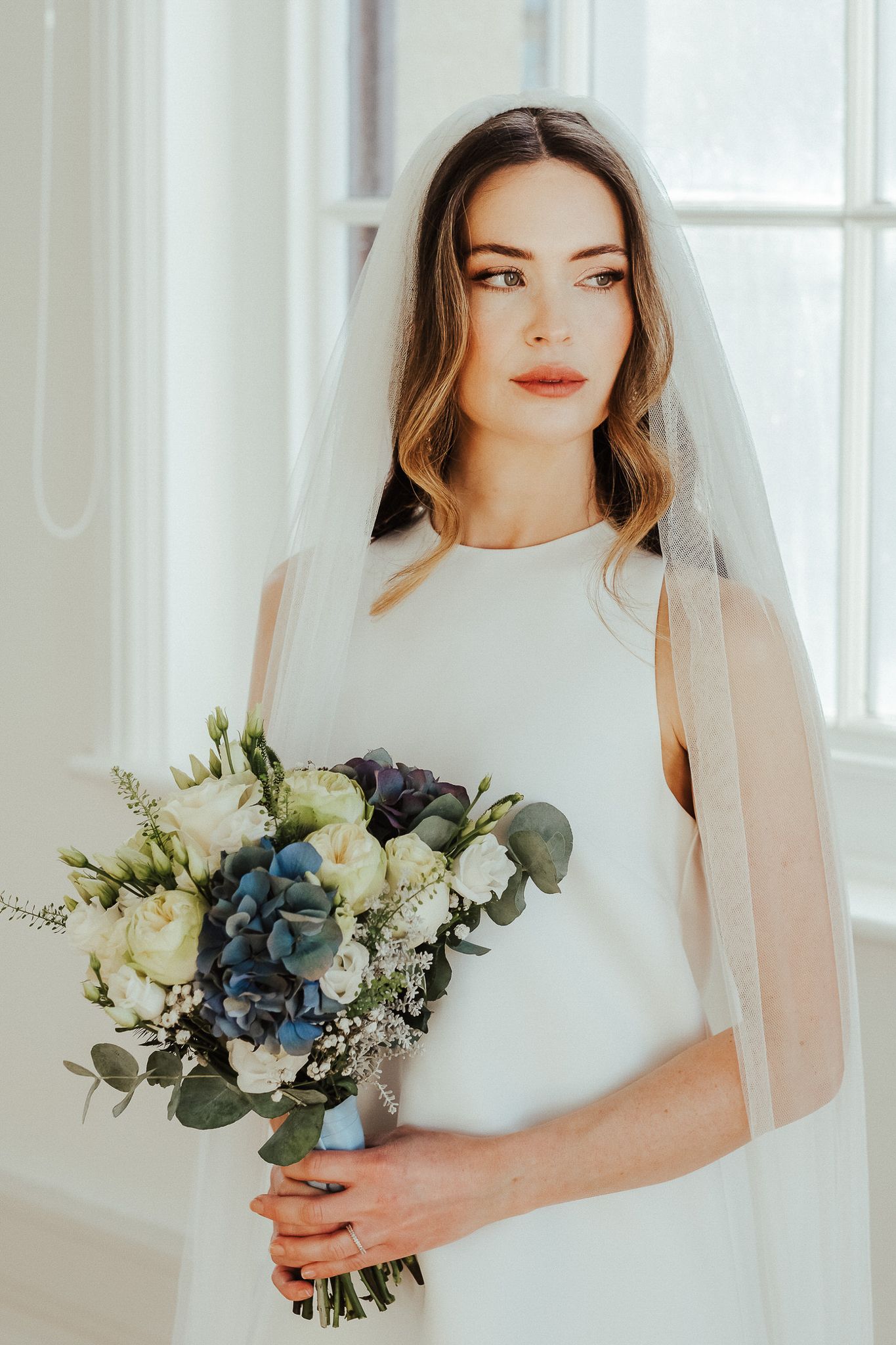 Bride in elegant white wedding dress holds blue and white floral bouquet in bright sunlit room.