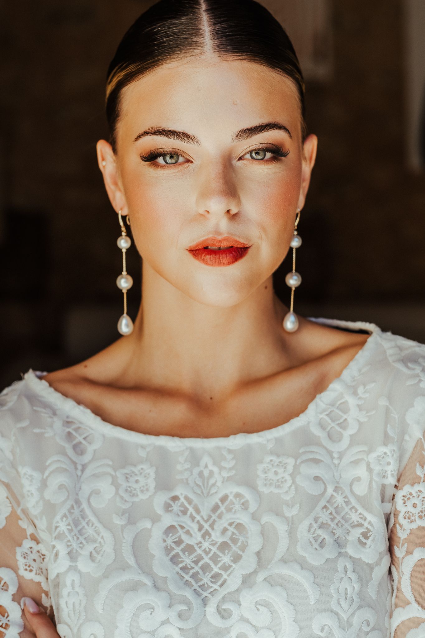Close-up portrait showing pearl drop earrings and white lace dress with bright orange-red lipstick.