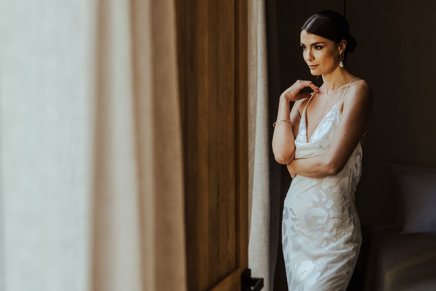 Bride in elegant white dress poses near window in moody natural light for wedding portraits.