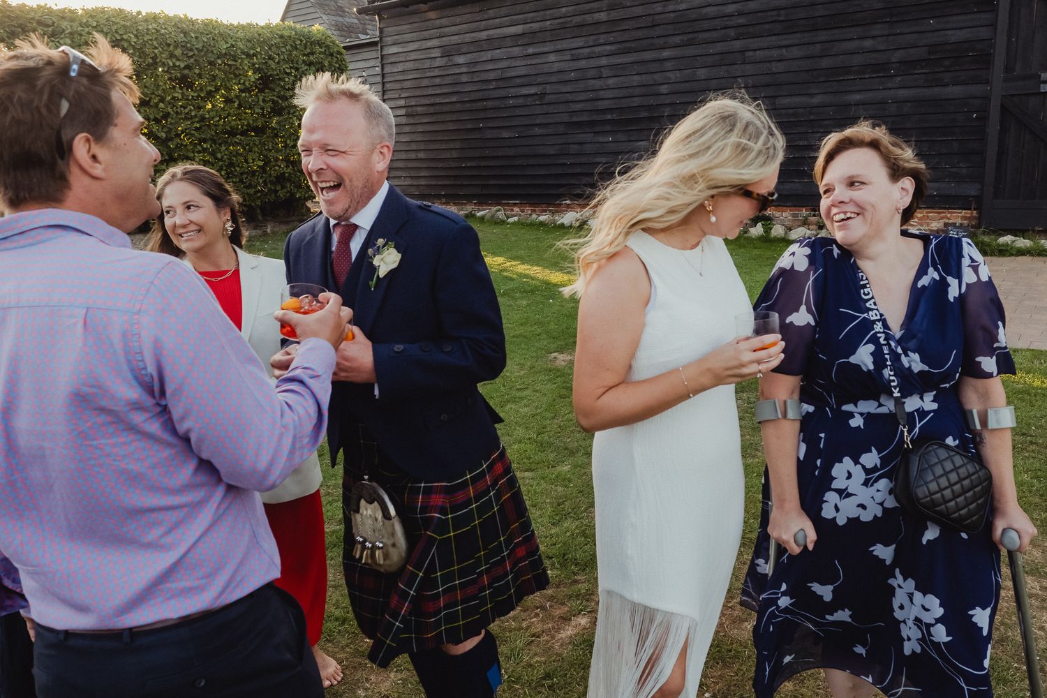 Wedding guests laugh and socialize outdoors, with one guest wearing a traditional Scottish kilt at a garden reception.