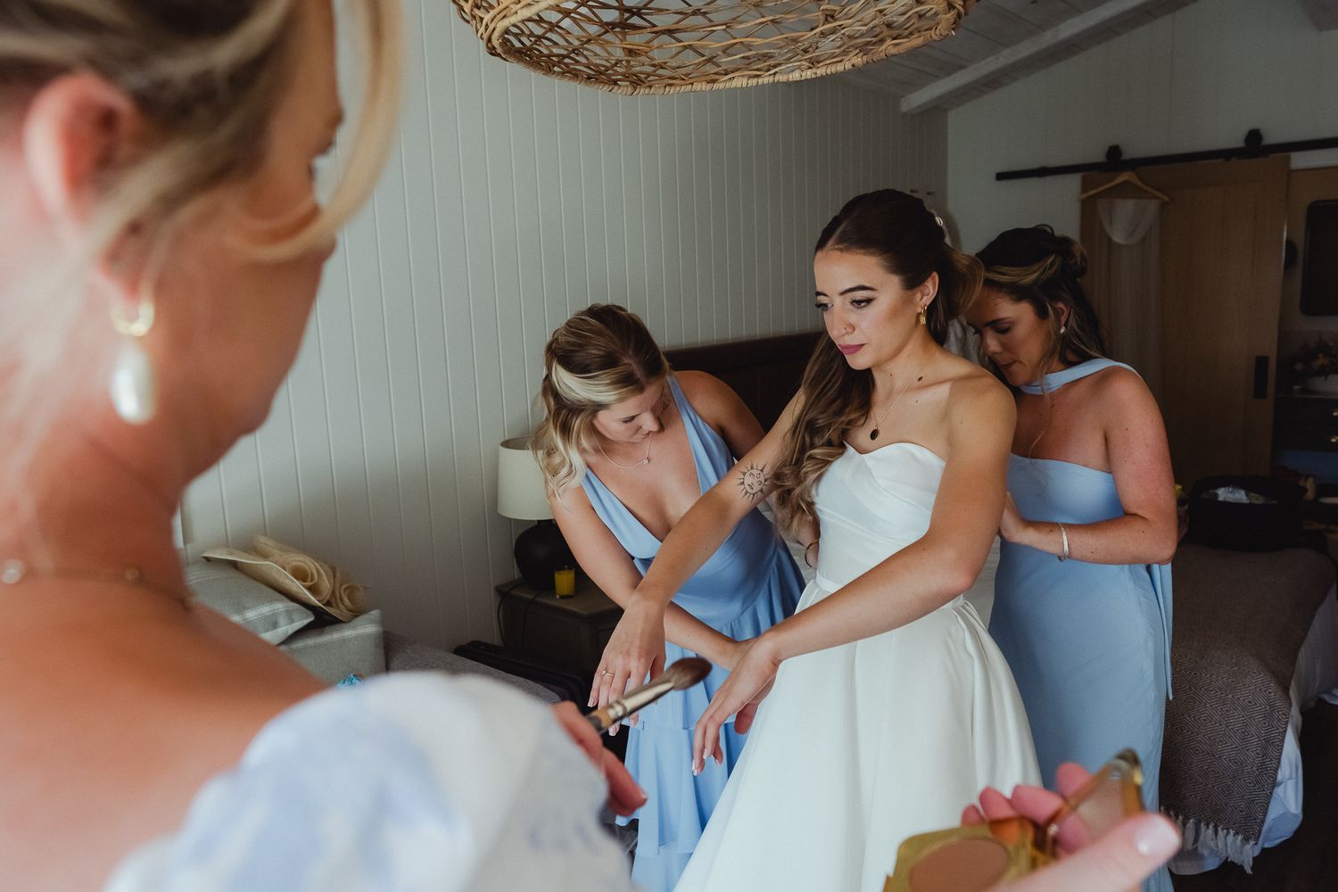 Bridesmaids in light blue dresses help the bride get ready for her wedding day in a sunlit room.