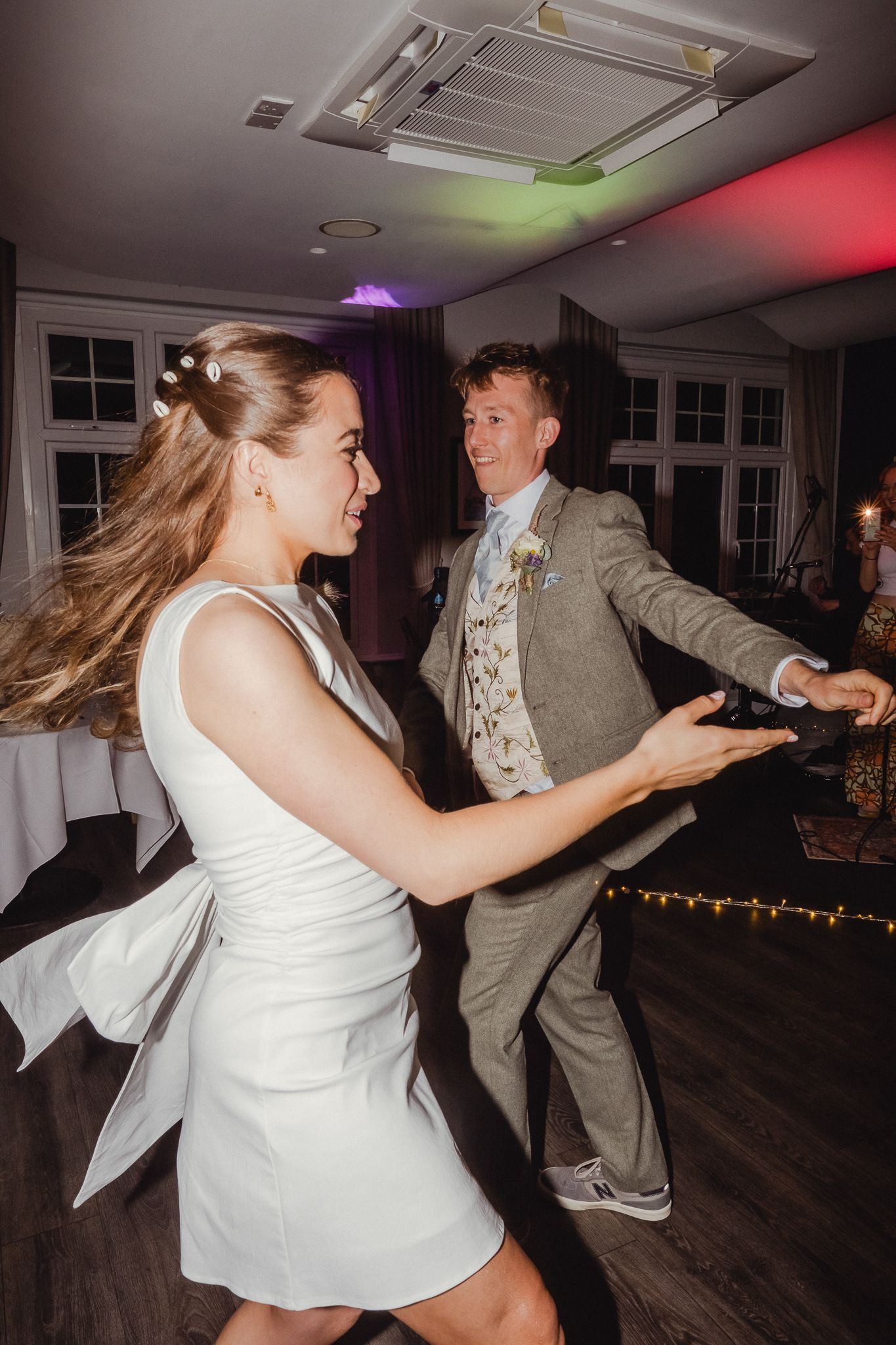 A couple dances energetically at a wedding reception with colorful lighting and decor visible in the background.