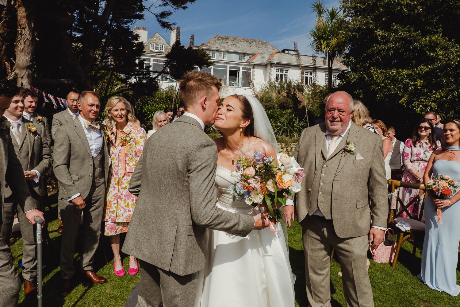Wedding party members in light gray suits and blue dresses gather outdoors for celebratory photos on a sunny day.