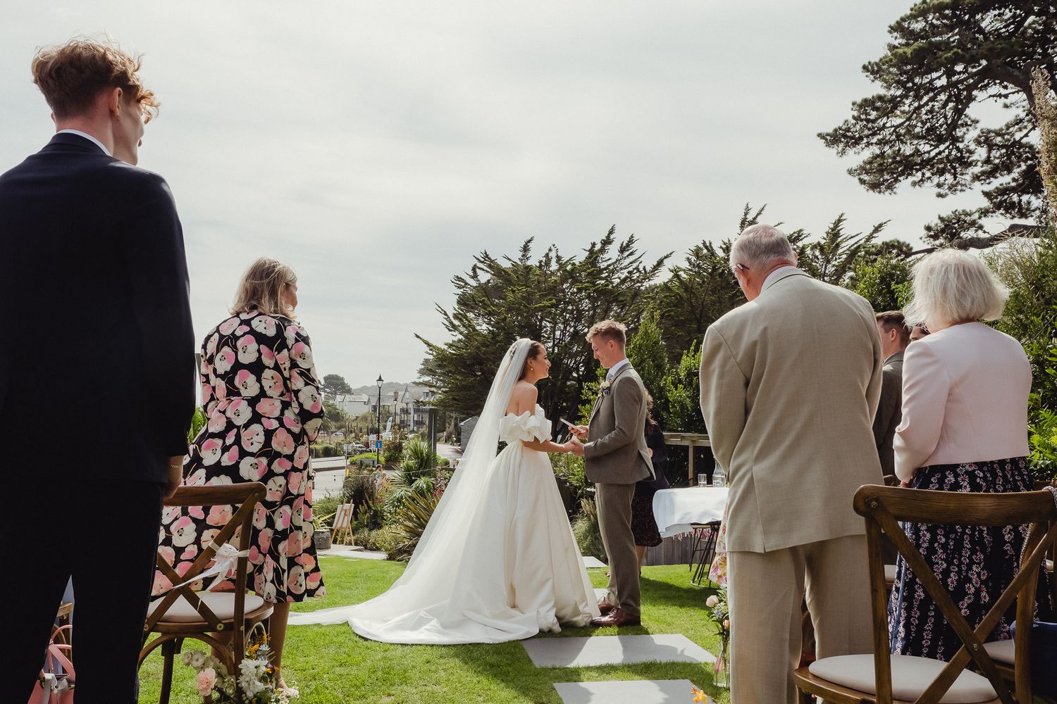 Wedding guests gather outdoors to witness a ceremony overlooking a scenic coastal landscape on a cloudy day.