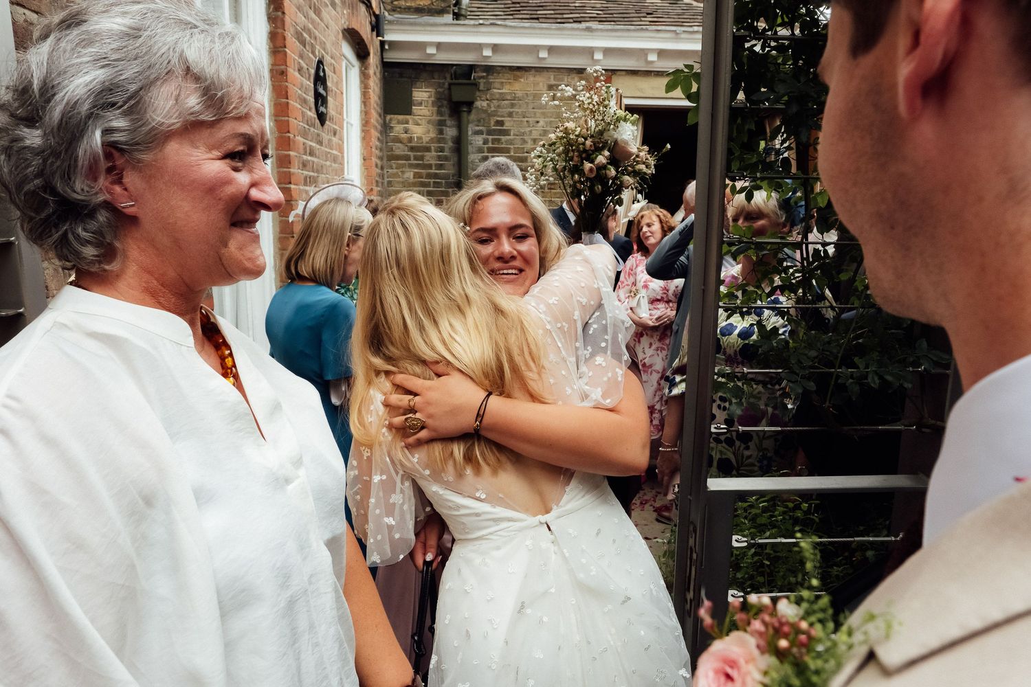 Bride in a white lace dress shares a warm embrace with a smiling older woman at an outdoor wedding celebration beside a brick building.