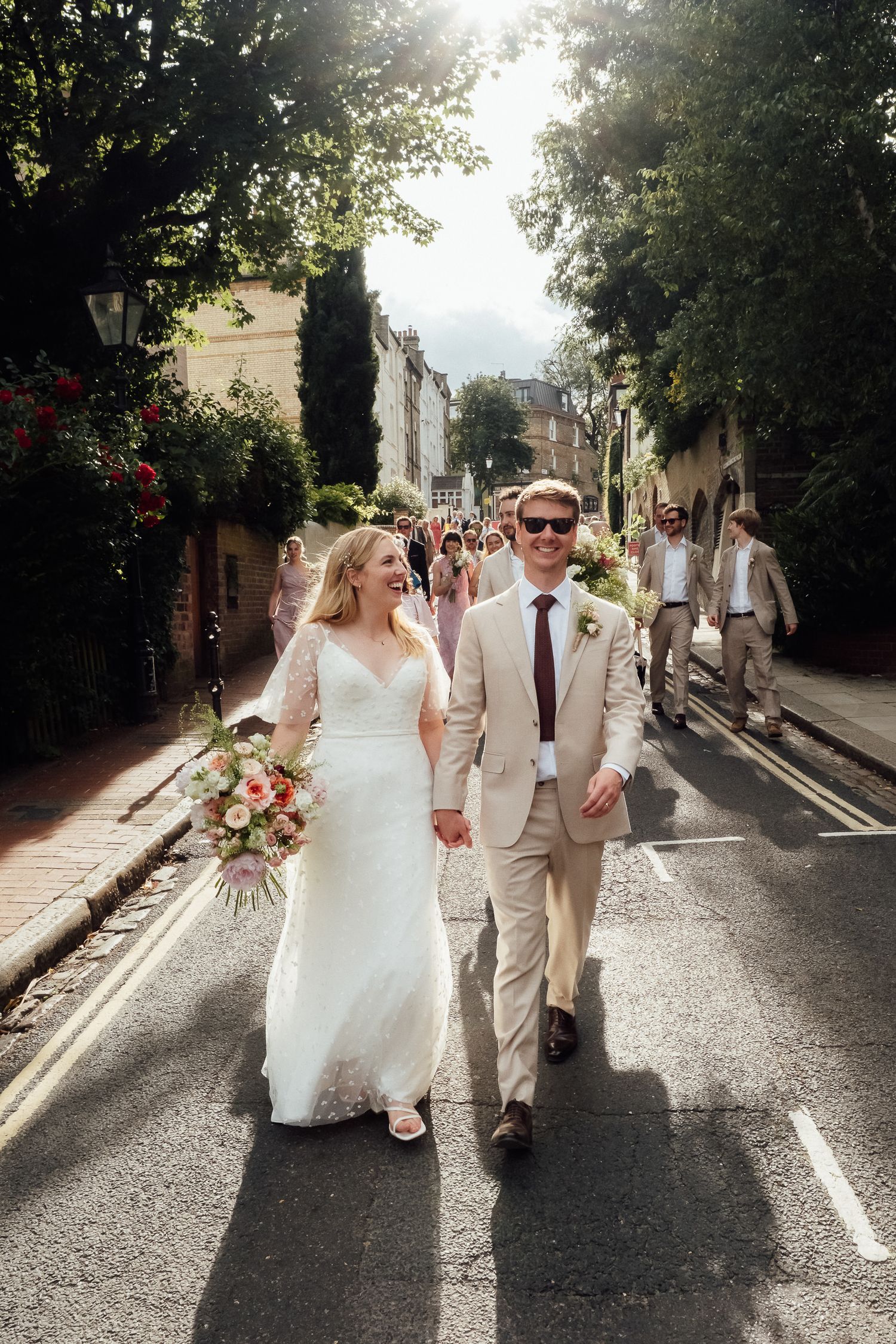 Smiling bride in a lace gown and groom in a tan suit walk hand in hand down a sunlit street, followed by wedding guests.