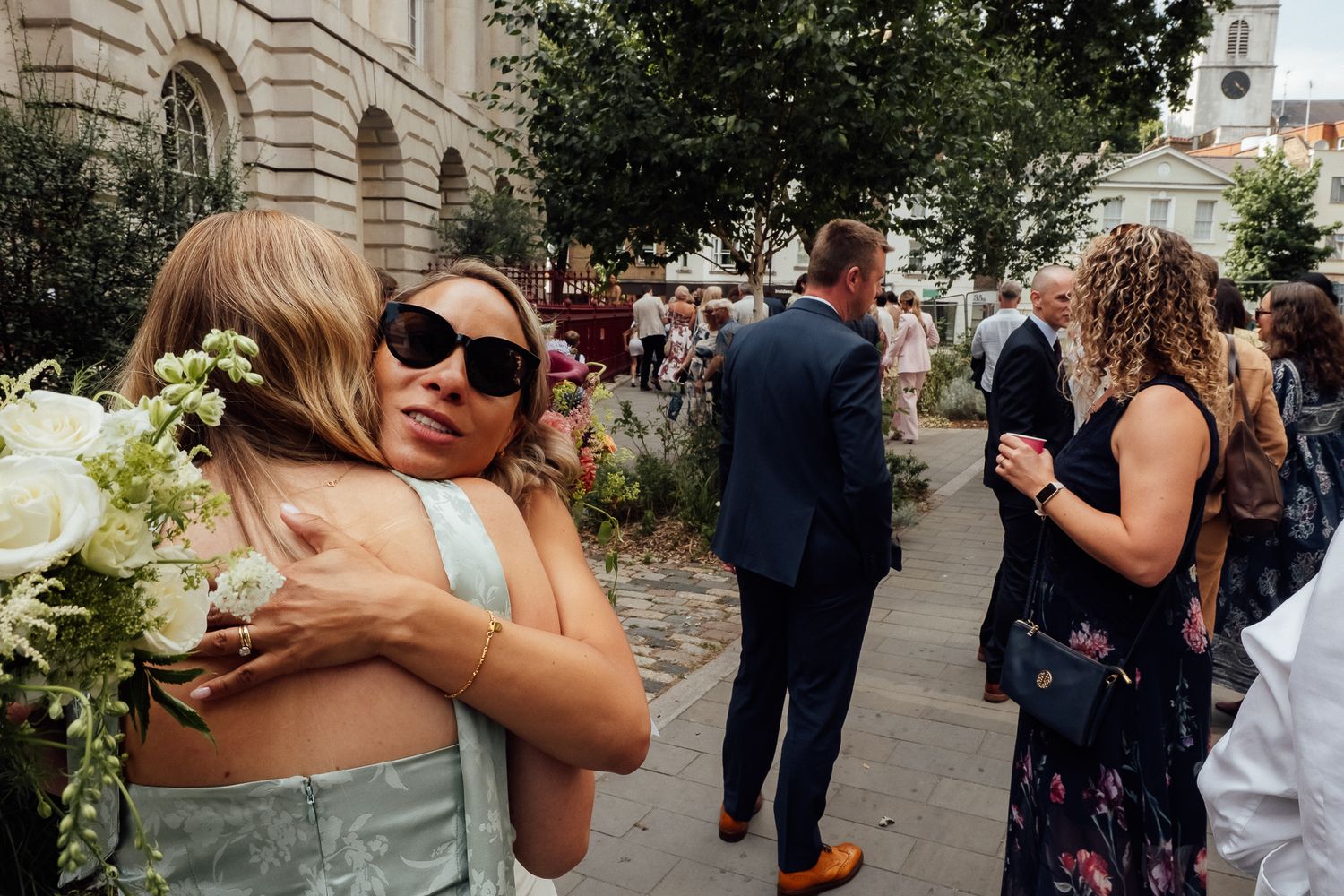 Woman in sunglasses and green dress hugs a guest holding white flowers outside a stone building as wedding guests gather behind them.