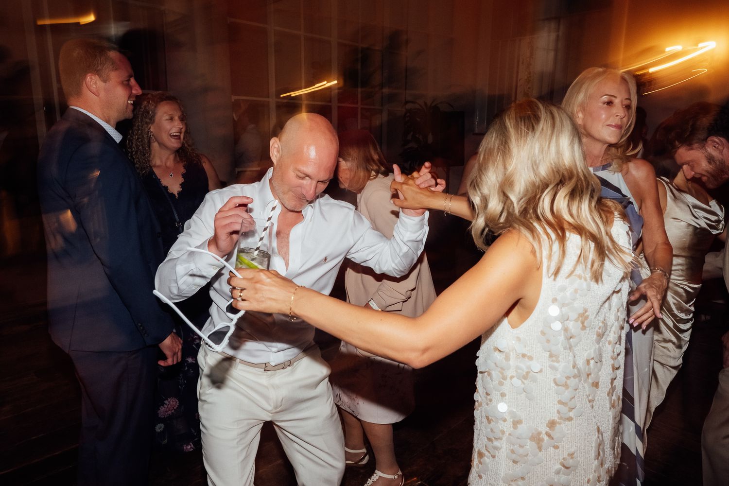 Wedding guests dance and laugh together on a warmly lit dance floor, with a bald man in a white shirt holding a drink.