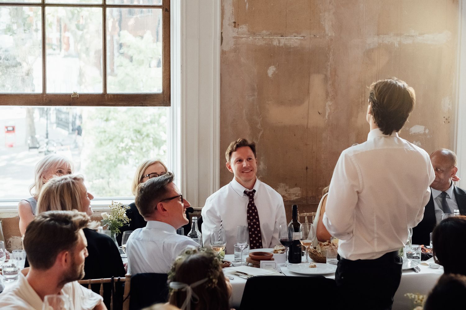 Man in a white shirt stands and addresses guests seated around a banquet table in a sunlit room with large windows.