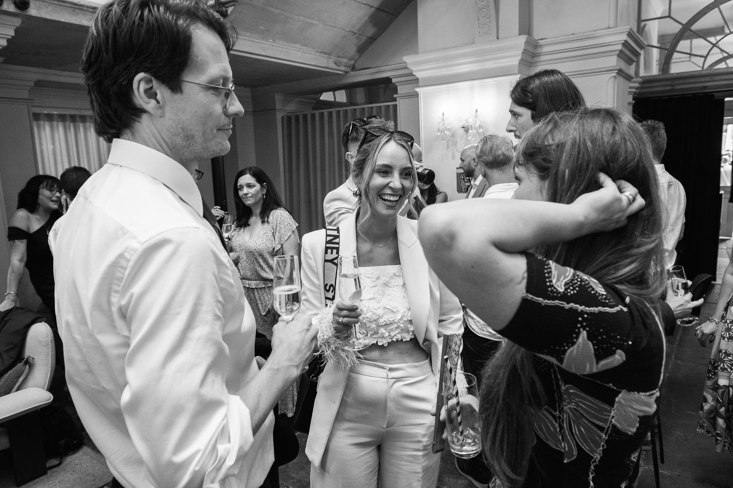 Smiling woman in a white outfit holds a drink while chatting with guests at a lively indoor party in black and white.