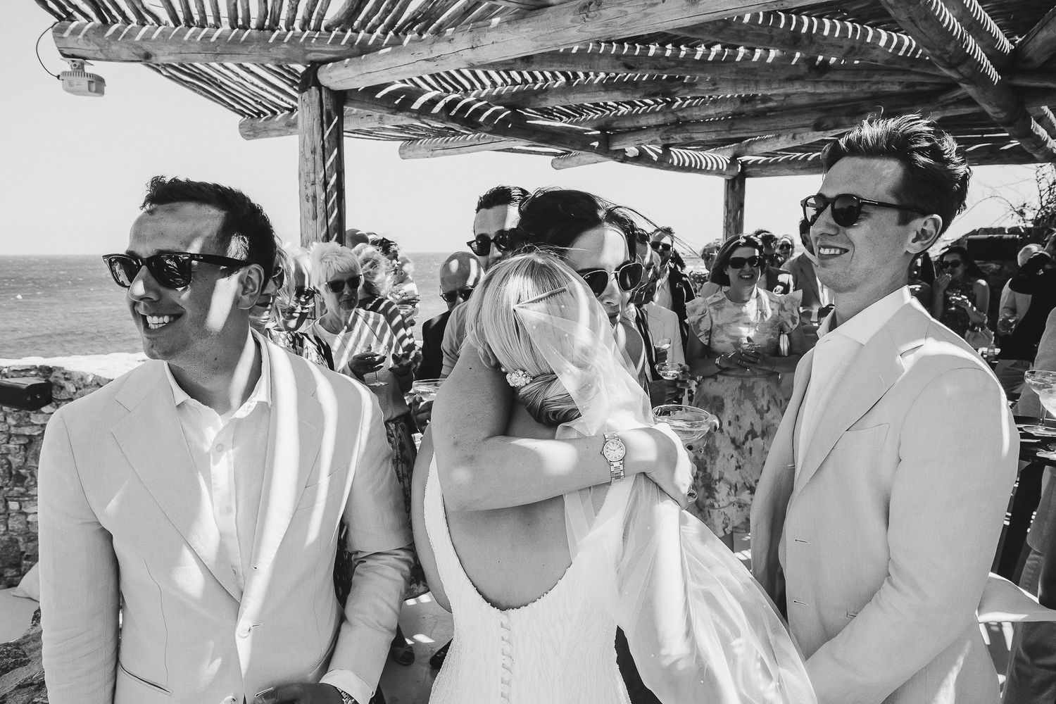 Group seated at outdoor restaurant wearing sunglasses and enjoying tropical drinks during vacation celebration.