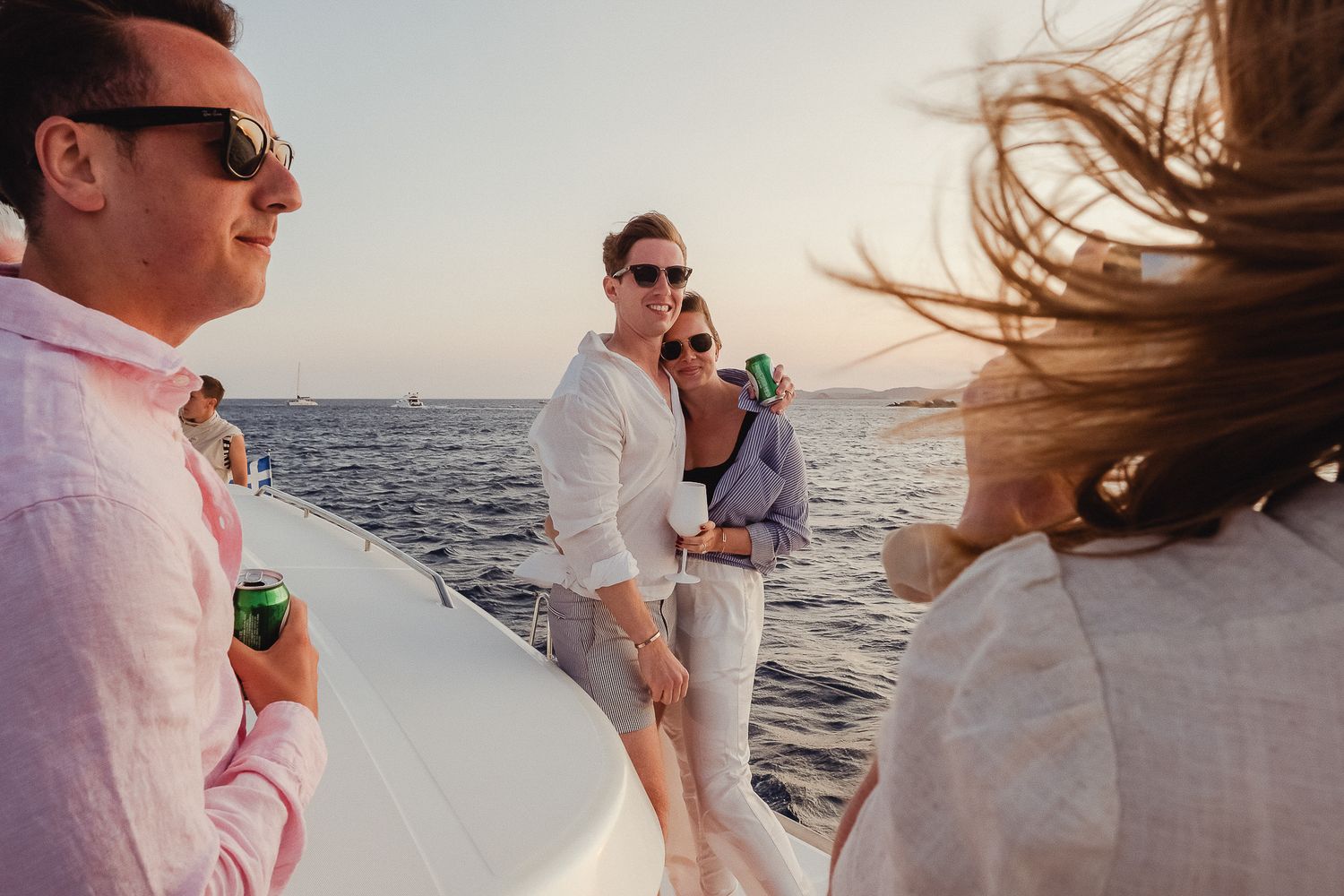 Wedding party celebrates on a sailboat at sunset with flowing dresses and champagne flutes against the ocean backdrop.