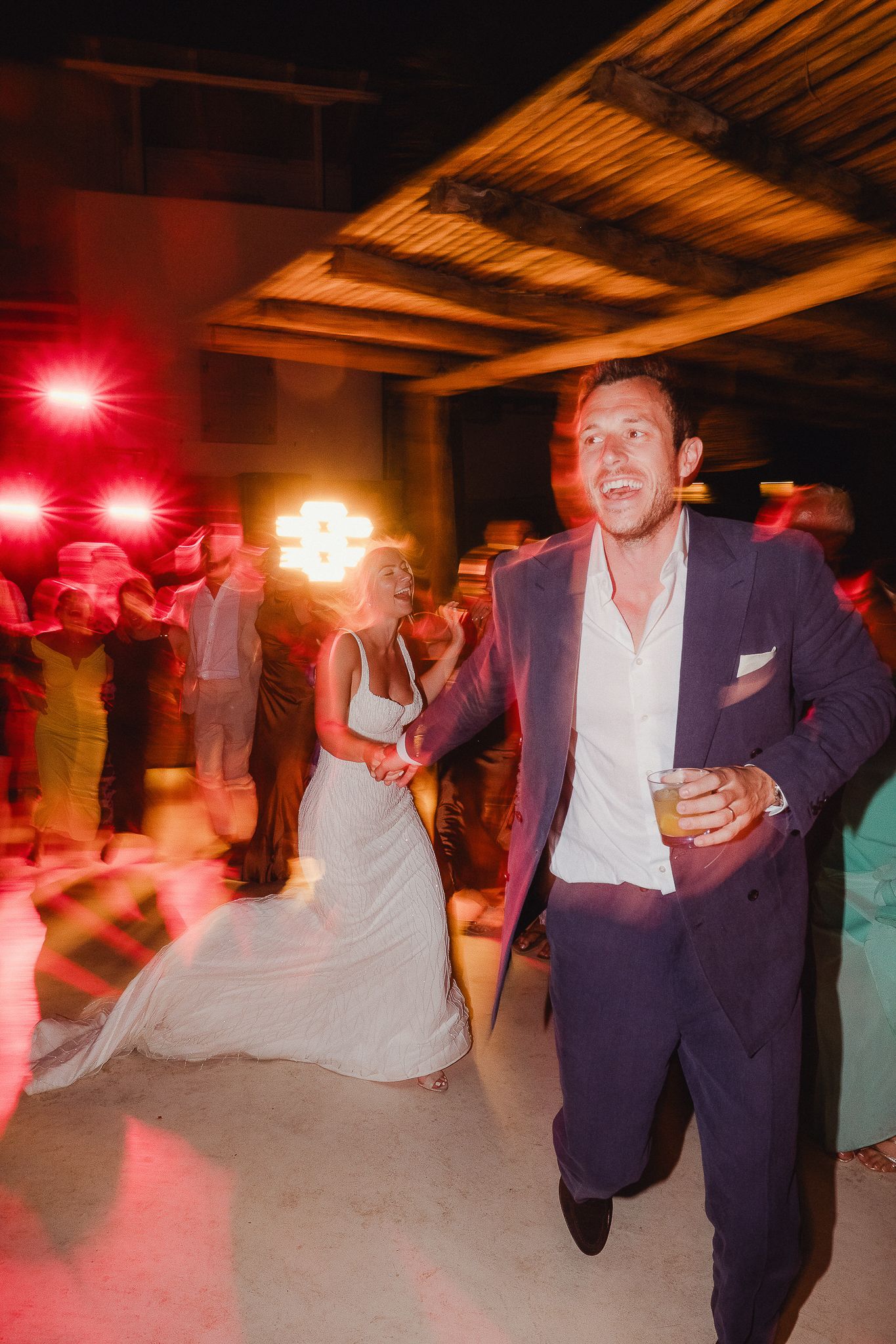 Dancers move energetically on a dimly lit dance floor illuminated by red and white spotlights during an evening celebration.