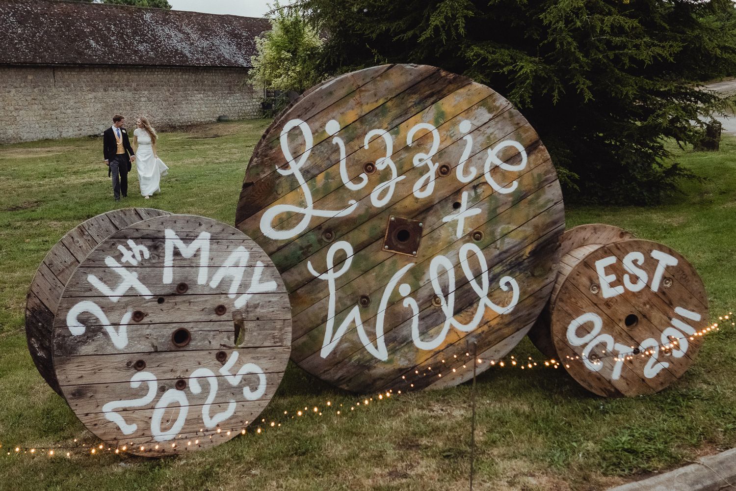 Rustic tree logs decorated with white painted writing and fairy lights line up against a stone wall in a garden area.