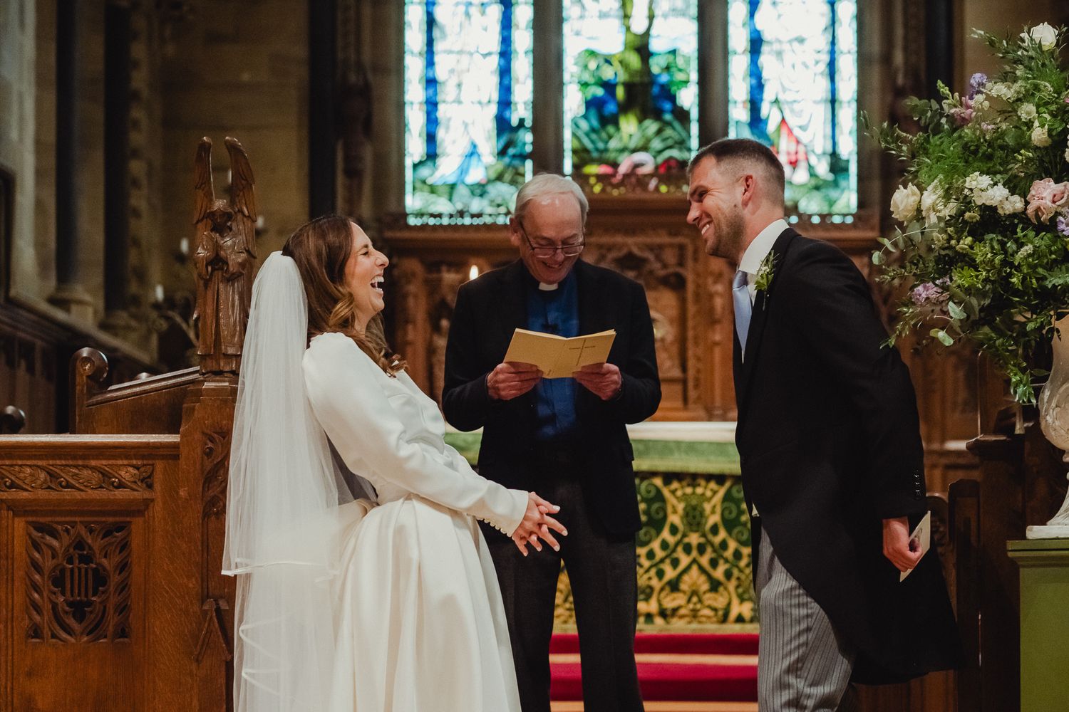 A couple exchanges vows during a traditional church wedding ceremony with stained glass windows and floral decorations.