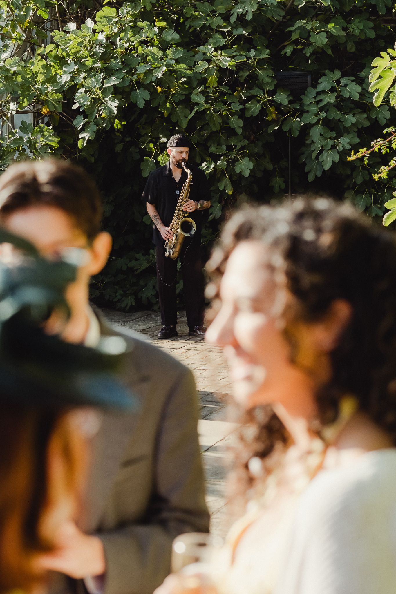 A saxophone player performs in an outdoor garden setting while guests enjoy the music.