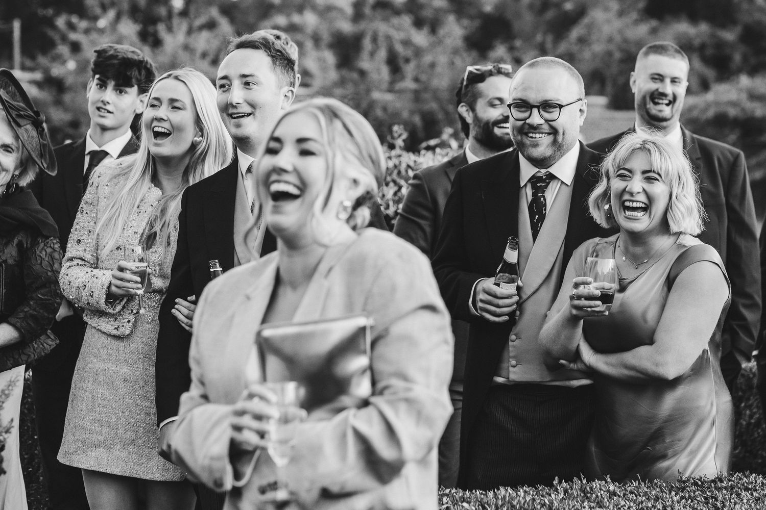 Black and white photo of wedding guests laughing and enjoying drinks during an outdoor celebration.
