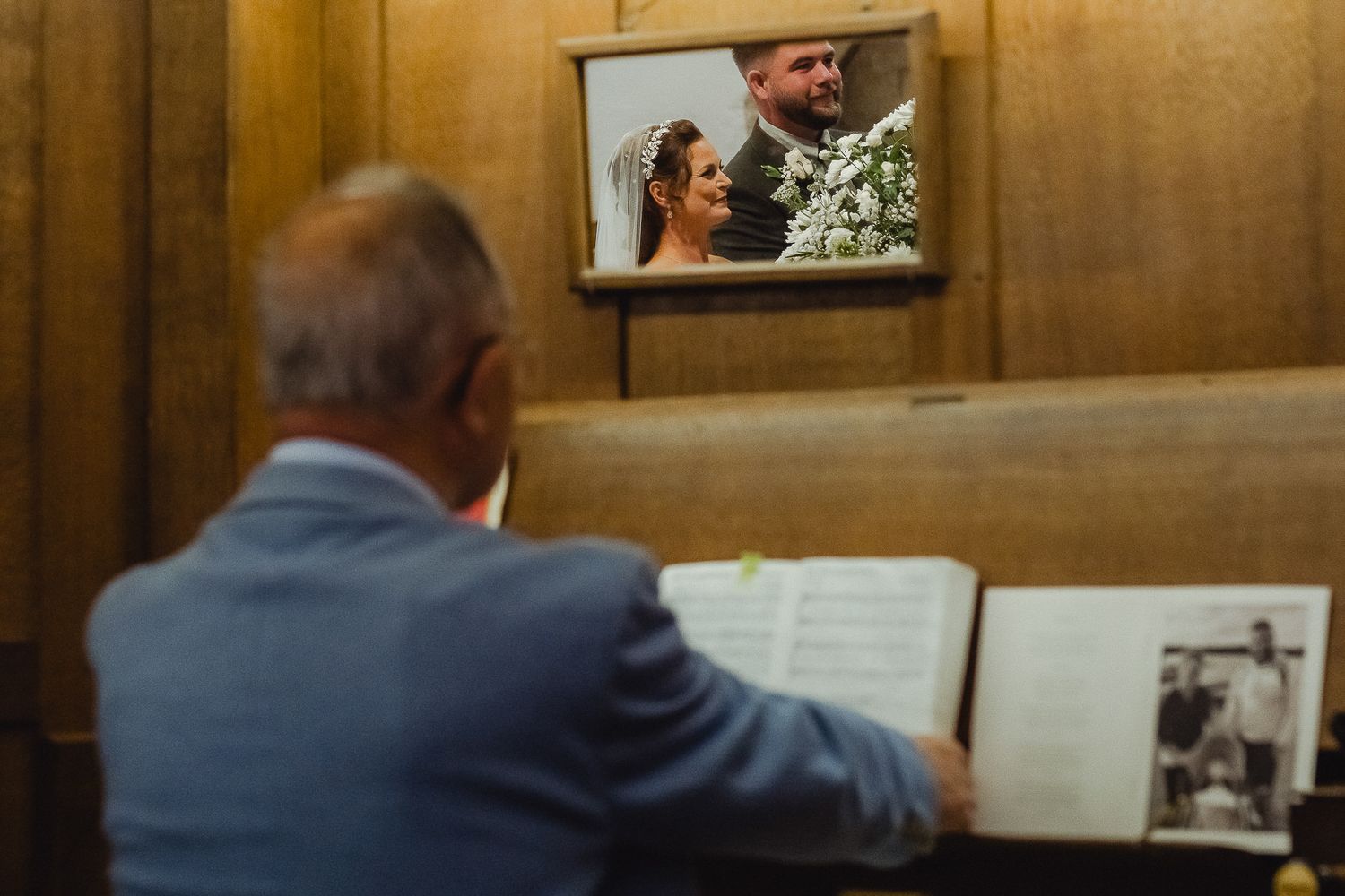 A person in a blue suit looks at a wedding photo album while a wedding scene plays on a mounted TV screen above.