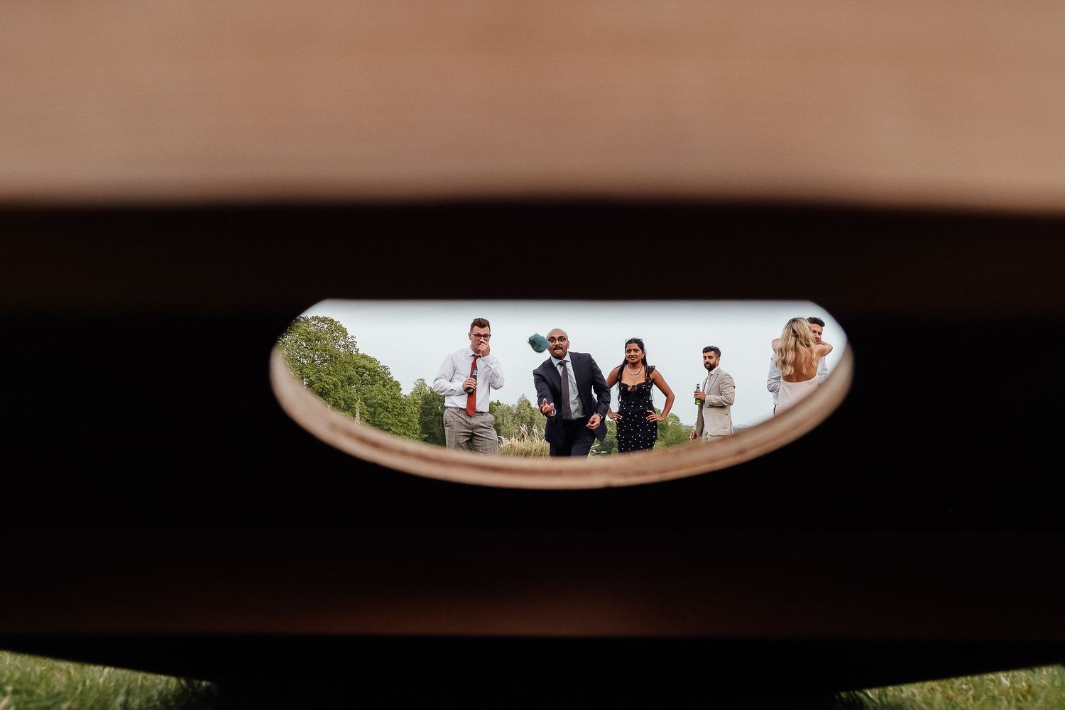 Wedding party viewed through a wooden bench cutout, creating a unique framed perspective of elegantly dressed guests outdoors.
