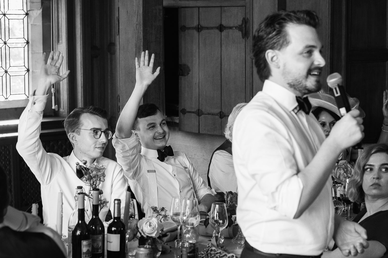 Men in formal attire celebrate at a wedding reception, one raising his hand while another speaks into a microphone.