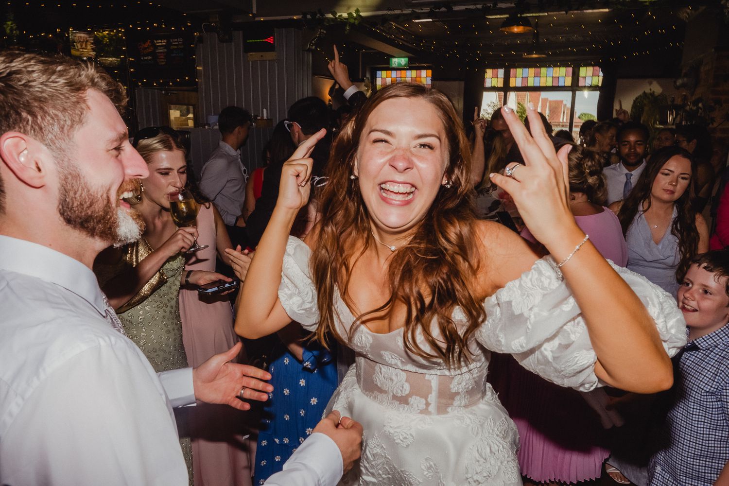 Wedding guests dance and celebrate on a crowded dance floor at an indoor reception venue during evening festivities.