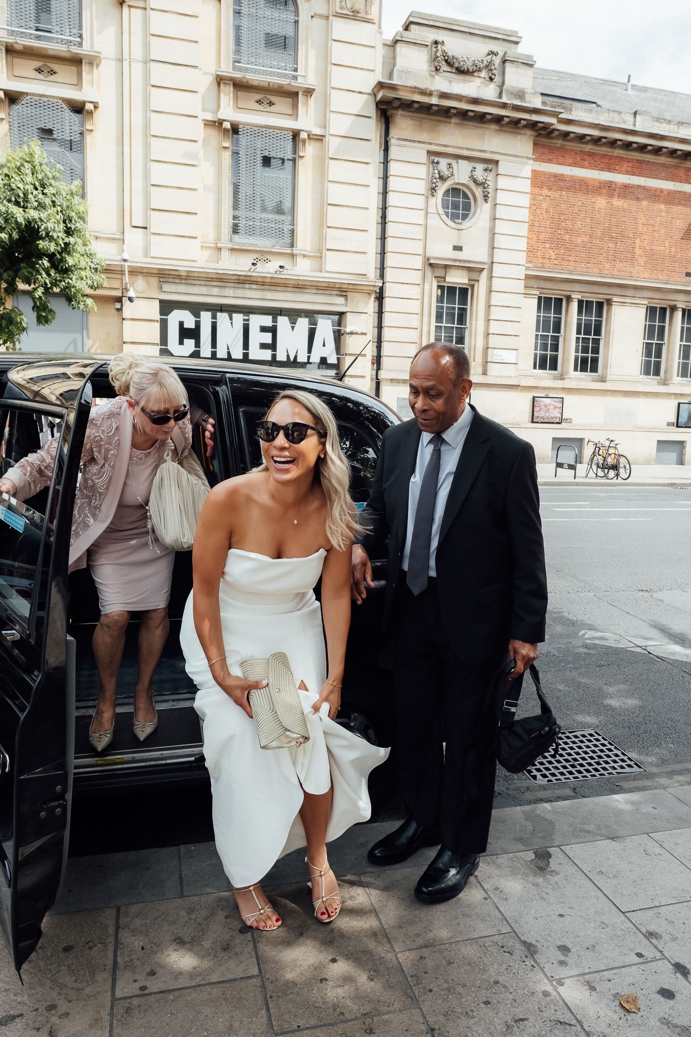 Smiling bride in a white strapless dress steps out of a black car outside a cinema, greeted by a man in a suit and a woman in sunglasses.