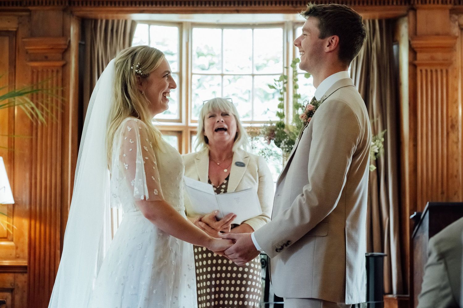 A smiling bride in a white lace gown and veil holds hands with her groom as a laughing officiant reads vows during their wedding ceremony.