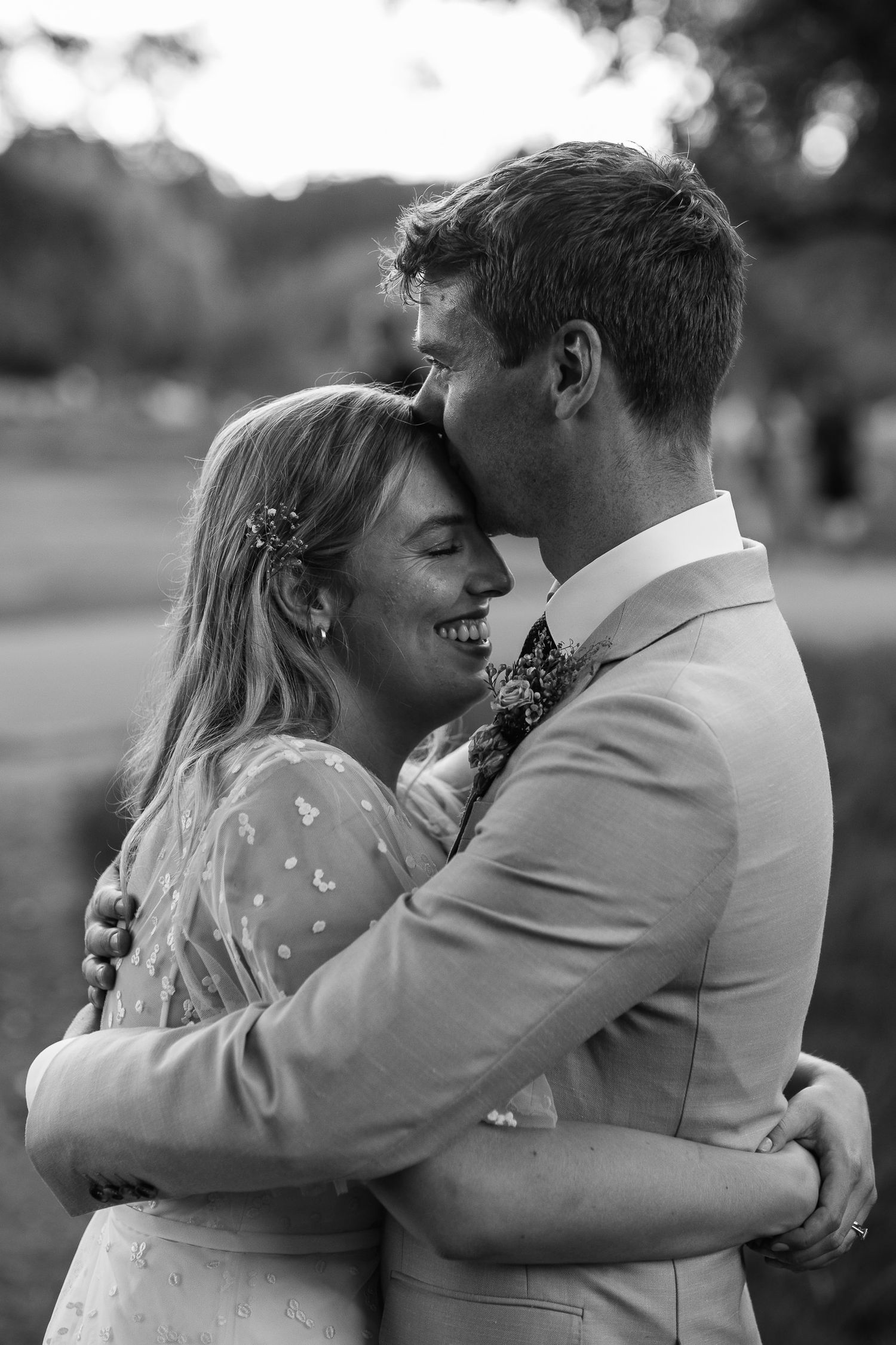 Black and white photo of a smiling bride with a floral hair clip embraced by a groom in a suit as he kisses her forehead outdoors.