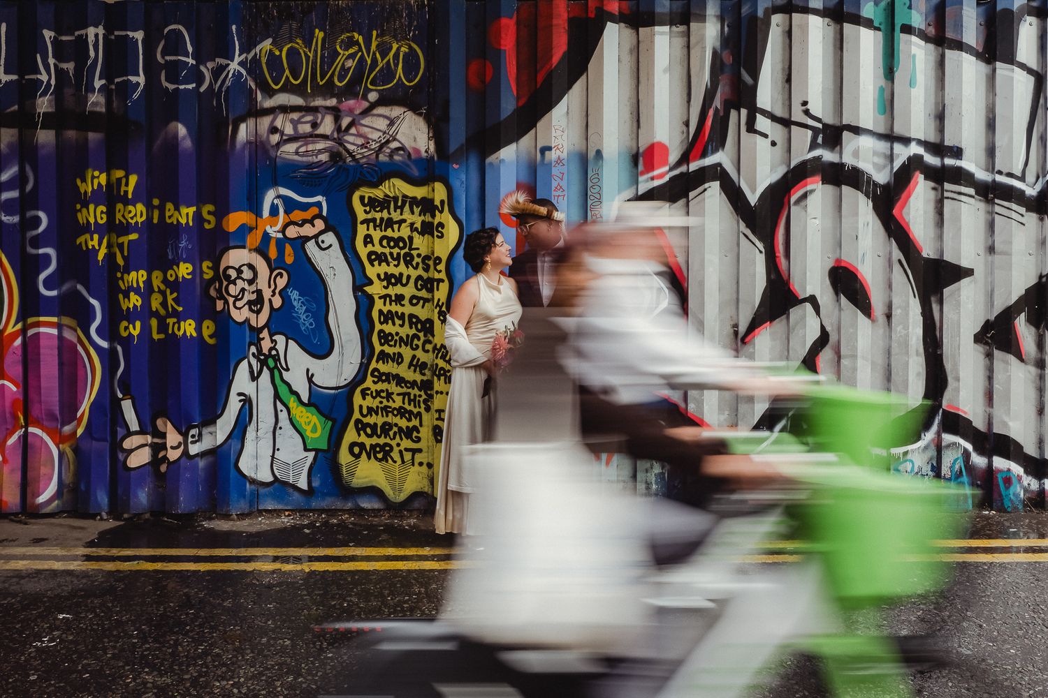 Blurred motion of cyclists riding past colorful graffiti murals on an urban wall.