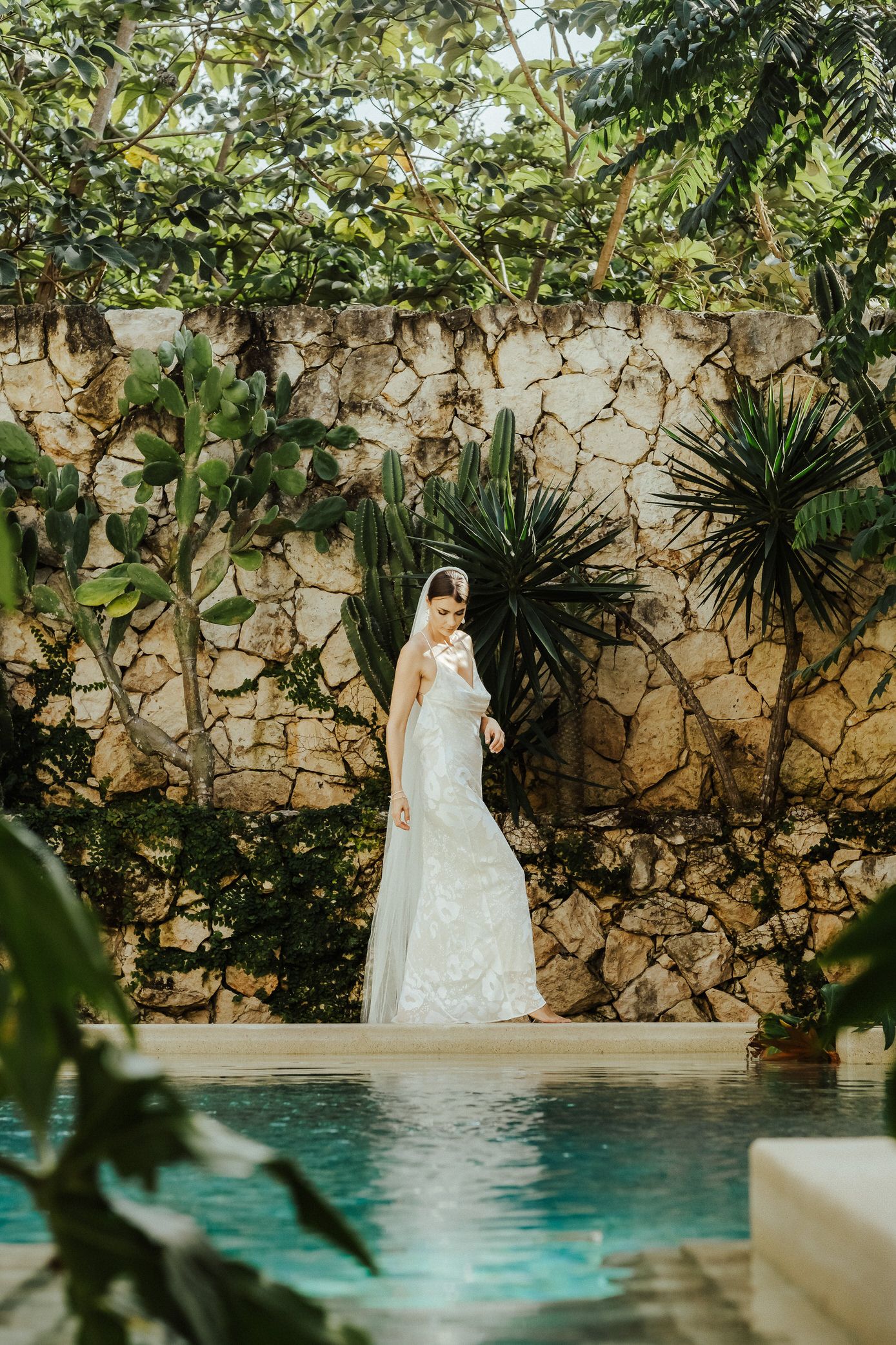 A white wedding dress stands elegantly against a stone wall beside a turquoise pool surrounded by tropical plants.