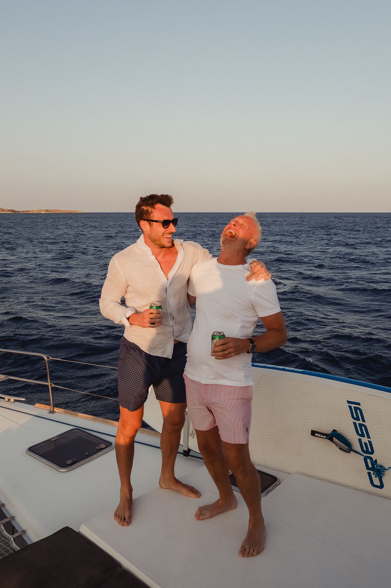 Two people laughing and enjoying drinks together on the deck of a boat during sunset at sea.