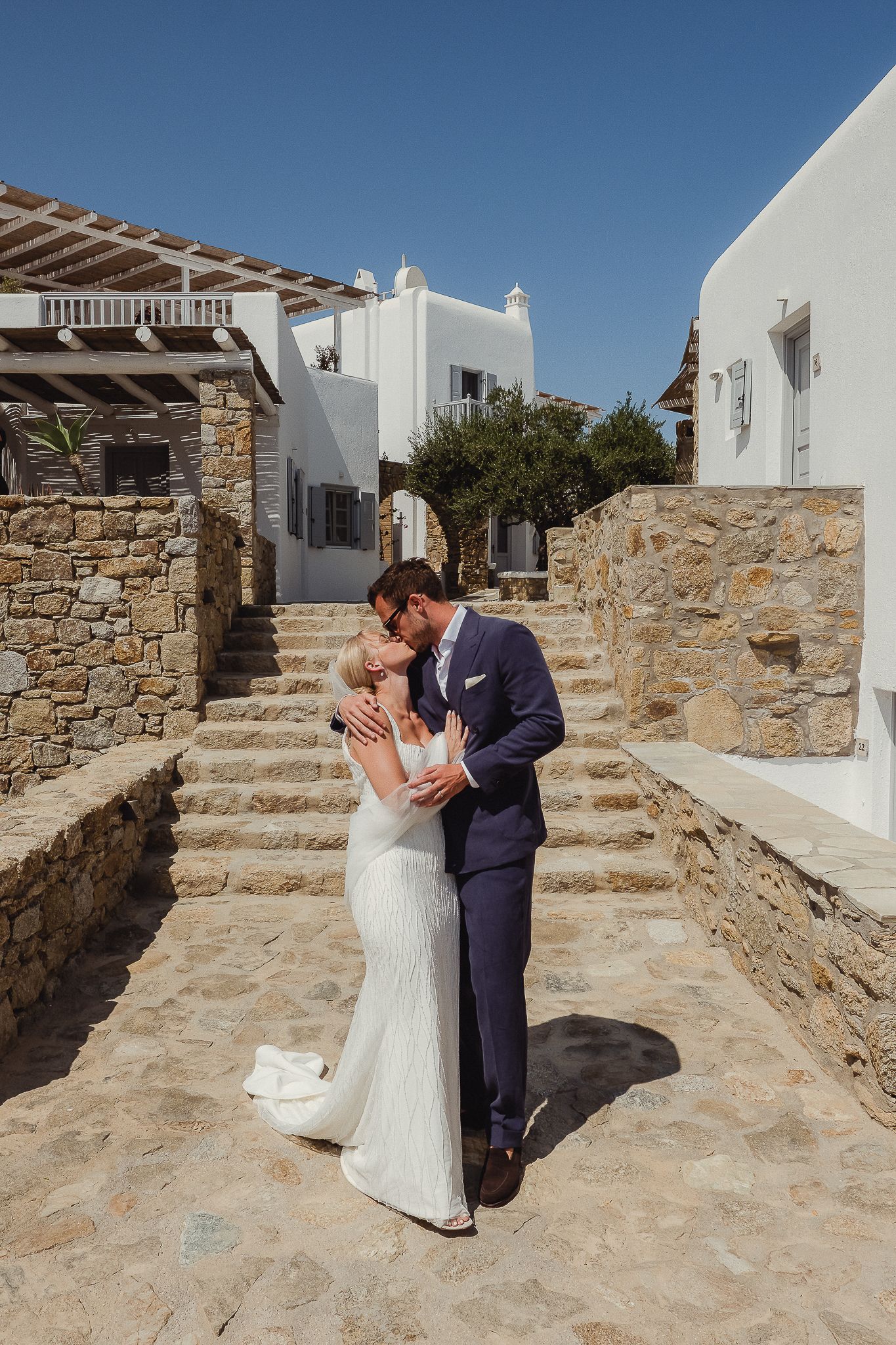 A couple shares a romantic first look moment on stone steps along a whitewashed Mediterranean villa wall on their wedding day.