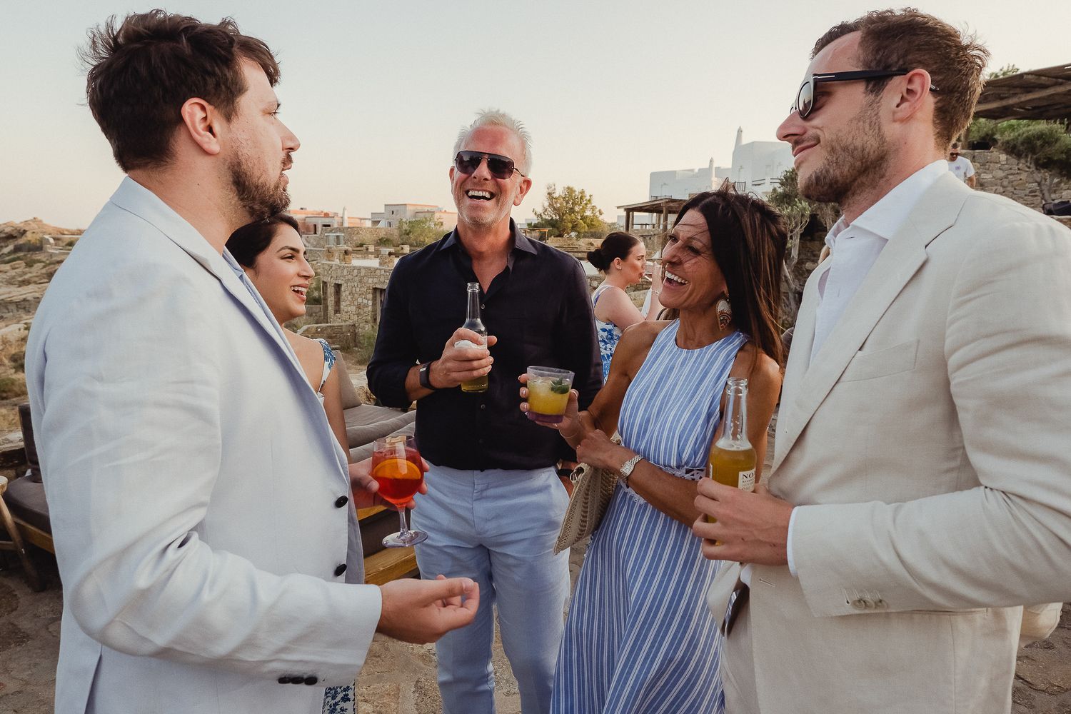 A group of people in light summer attire attend a rooftop cocktail party overlooking a city.